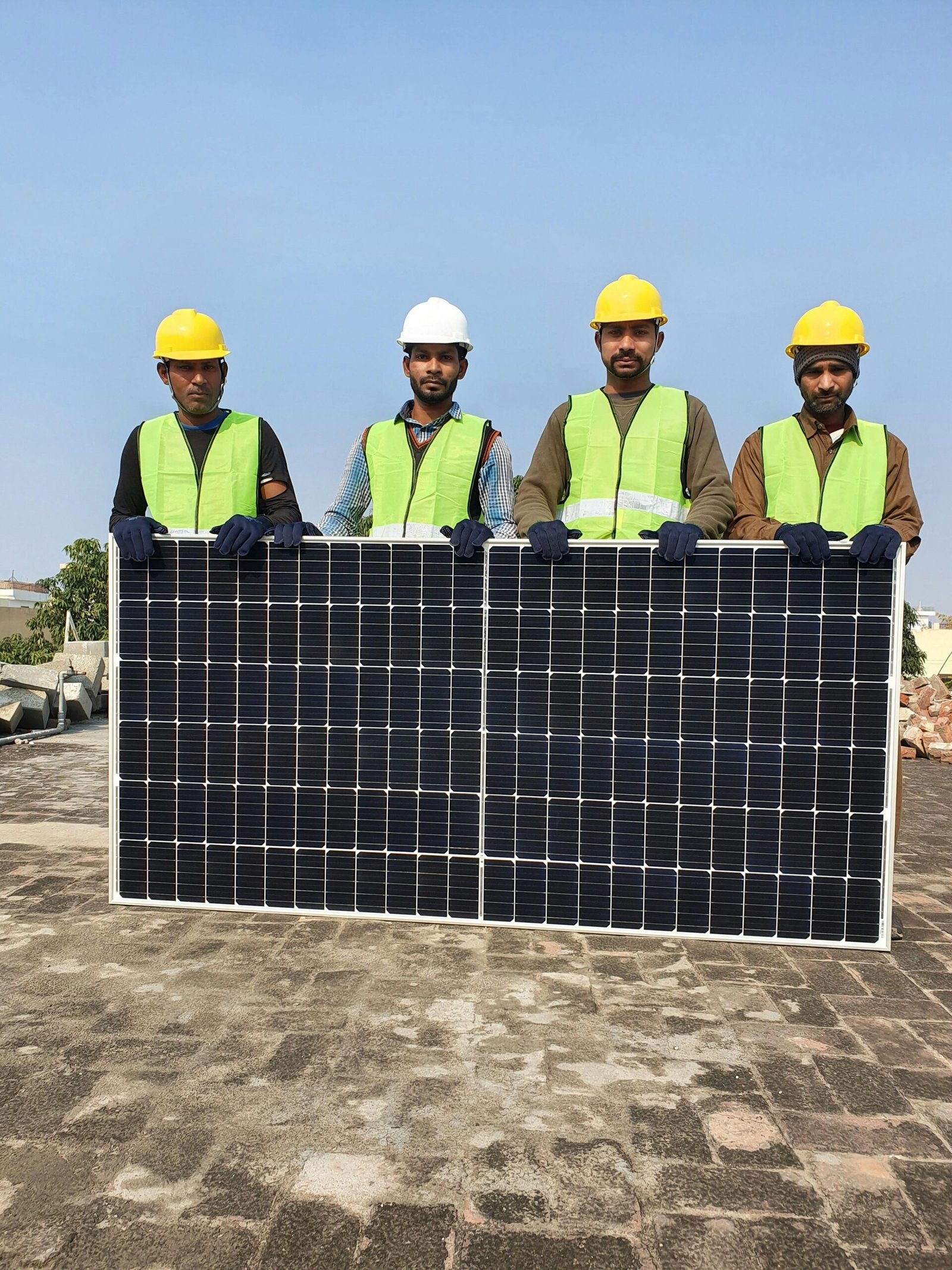 Group of construction workers holding a solar panel on a sunny day, wearing safety gear.